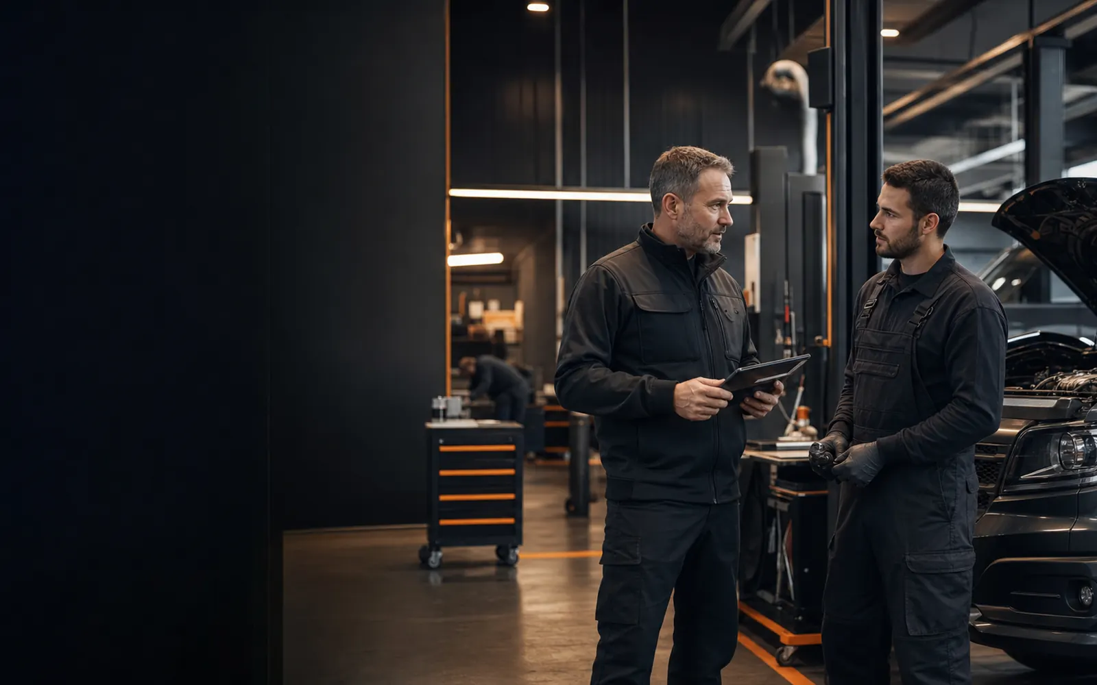 Workshop manager and mechanic discussing technical work beside a vehicle.
